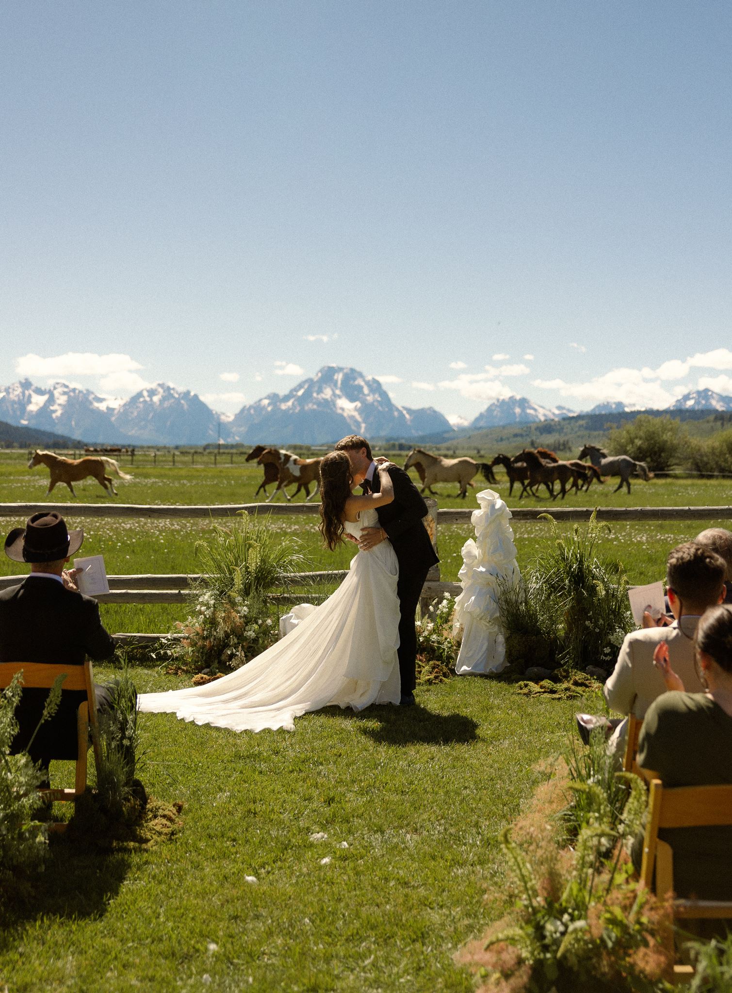 Bride and groom embracing with horses running behind them in a mountain meadow